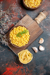 Above view of uncooked pastas in a brown bowl and garlic on wooden cutting board on mixed color background