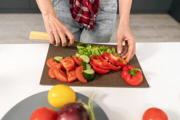 Obraz na płótnie Canvas Cutting a fresh vegetable salad young housewife cut tomato, cucumber, pepper on board preparing for a family dinner standing in the kitchen. Healthy food living. Healthy lifestyle. 