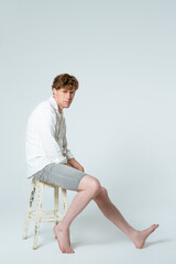 Vertical image. Sitting half turned on the rusty chair full length shot of young handsome young man wearing white shirt and grey shorts looking at camera isolated on white background. 