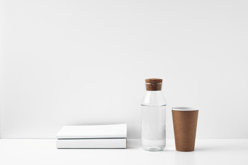 Transparent bottle with cork stopper paper cup and books on a white background. Natural and eco-friendly materials in interior decor. Copy space, mock up.