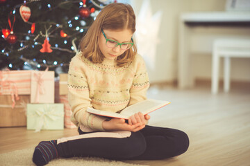 Portrait of pretty child girl reading book sitting near christmas tree