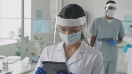 Chest up shot of Asian female doctor in lab coat, face shield and mask using digital tablet while working in medical office during covid-19 outbreak