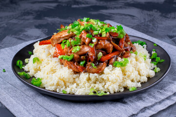Stir fried turkey pieces with mushroom, carrot and garlic on top of couscous. Appetizing hot meal garnished with green onion served on the grey table