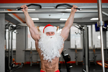 A man wearing a santa claus hat and a naked torso pulls up on a horizontal bar in a gym.