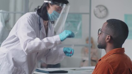 Female doctor in protective face screen, mask and gloves collecting nasal swab from young Afro-American man while testing him for coronavirus in medical office - Powered by Adobe