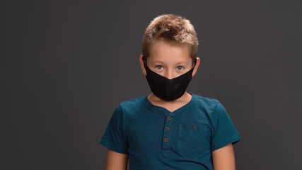 School boy in black reusable face mask wearing dark blue button neck t-shirt surprised look at camera while standing in studio. Isolated on black background. Kids fashion concept. 