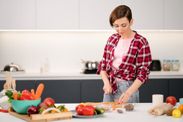Obraz na płótnie Canvas Cutting fresh carrot housewife cooking stew meat or ragout wearing a plaid shirt. Cooking with passion young woman with short hair standing at modern kitchen. Healthy food leaving - concept. 