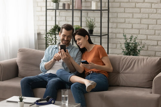 Sharing information. Happy loving young spouses sitting on couch at living room using smartphones exchanging files. Couple of diverse friends showing each other funny photo video at social network
