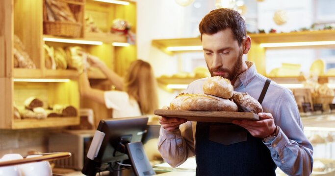 Close up of happy bearded young Caucasian man seller in apron standing in bakery and holding tray with fresh baked bread in hands, smelling baking and looking at camera while colleague works behind