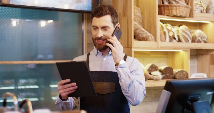 Portrait Of Happy Caucasian Bearded Handsome Male Baker Standing In Own Bakery, Tapping On Tablet Device And Speaking On Smartphone. Male Worker Talking On Mobile Phone With Client. Bakehouse Concept