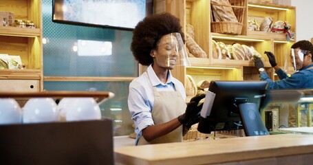 Close up of joyful smiling African American beautiful woman in apron using tablet tapping and typing while standing in bakery shop talking to her man coworker. Bakehouse concept