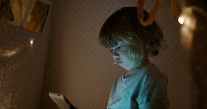 Cute funny caucasian baby boy is watching cartoons on a tablet while sitting in cozy tent with string lights 4k footage
