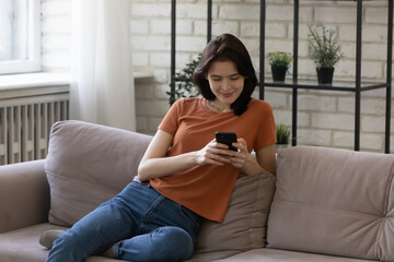 Love gives you wings. Happy young woman spend time sitting on couch at home chatting online with boyfriend. Millennial female texting tender message to beloved man enjoy reading email on phone screen