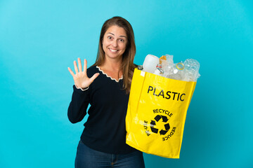 Middle age Brazilian woman holding a bag full of plastic bottles to recycle on blue background counting five with fingers