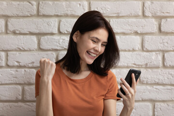 My victory. Excited young female standing by wall holding phone feeling satisfied of perfect exam result. Millennial woman cannot hold emotions reading good news from cell screen reaching success