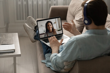 Glad to see you. Over the shoulder view of peaceful millennial man relaxing on sofa in earphone set with computer on laps. Calm young husband chatting with wife by video link using headphones and pc