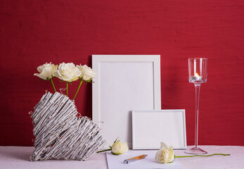 burning candle, writing materials, white roses in a vase, white photo frames, wooden heart on a red background