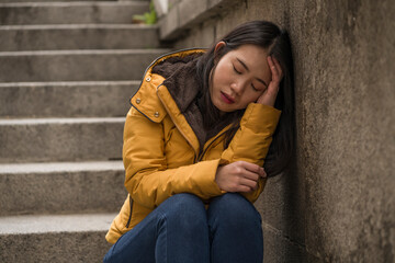 dramatic lifestyle portrait of young attractive sad and depressed Korean woman in winter jacket sitting outdoors on street corner staircase suffering depression problem