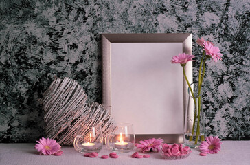 pink gerberas in a transparent vase and on the table, burning candles, photo frame and heart-shaped decor on a gray background