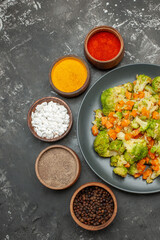 Vertical view of healthy meal with brocoli and carrots on a black plate and spices on gray table