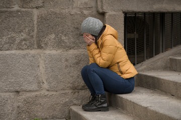 dramatic lifestyle portrait of young attractive sad and depressed Chinese woman in winter hat sitting outdoors on street corner staircase suffering depression problem crying
