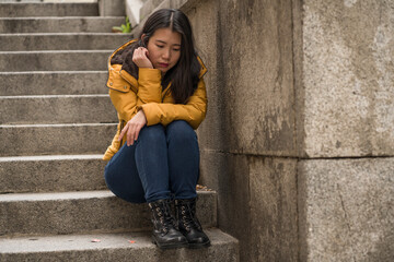 dramatic lifestyle portrait of young attractive sad and depressed Japanese woman in winter jacket sitting outdoors on street corner staircase suffering depression problem feeling helpless