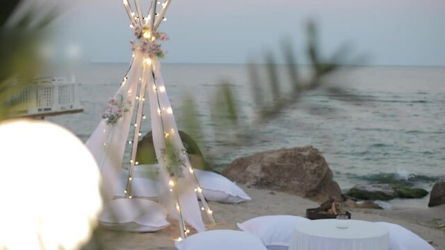 Lounge area of white pillows and tables on the seashore near the stones. The decorative tent is covered with a veil and hung with a garland.