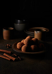 Chocolate truffles in cocoa dusting on dark brown ceramic plate. Dark brown ceramic plates and cups on background. Cinnamon sticks and star anise on the foreground. Low light moody photography