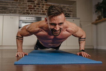 Fototapeta na wymiar A muscular man is doing pushups on a blue yoga mat in his apartment in the evening. An athletic guy with tattoos on his forearms is doing a chest workout at home. Bodybuilder with a naked torso