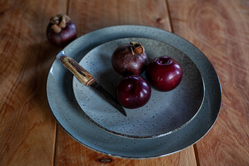 Whole purple plums and mangosteens and knife on ceramic plate on wooden table