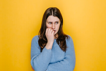 Worried young woman folded hands together near mouth, gnawing nails, female feels uncertain, stressed. Confused brunette girl with frighten or panic gesture, isolated on yellow studio background