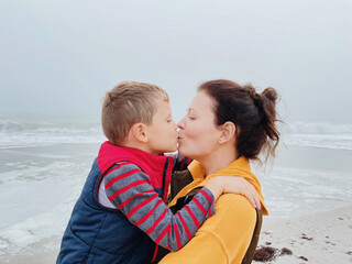 Fototapeta na wymiar Mother and son walk along the foggy beach and take a selfie on the phone. Portrait of happy mom kiss her child boy on the background of cloudy weather made with phone. Family time together. 