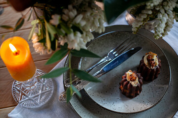 chocolate muffins in ceramic plates with fork and knife, yellow burning candle, textile napkin and flowers on wooden table