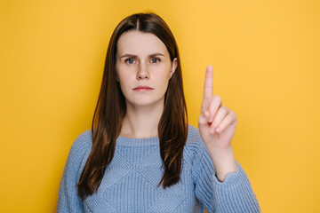 Portrait of young brunette woman warning with admonishing finger gesture, saying no, giving advice to avoid danger, dressed in blue sweater, disapproval sign, isolated on yellow studio background