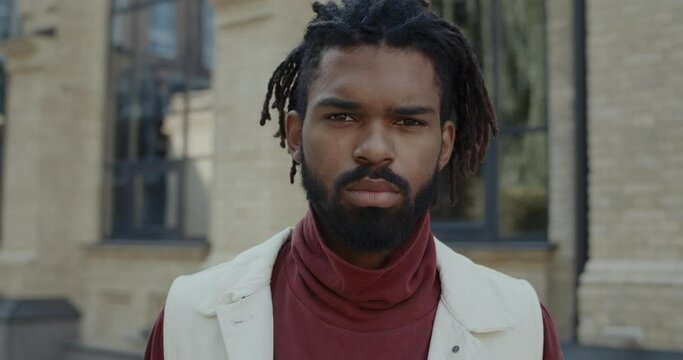 Close up view of bearded serious man turning head and looking to camera while standing at city street. Headshot of serious mixed race man with dreadlocks in turtleneck posing outdoors