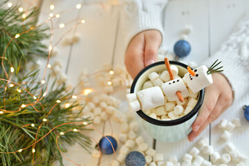 Girl holding cup of hot drink with marshmallow snowman at wooden table, top view. Bokeh effect. Happy New Year