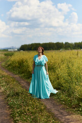 Fototapeta premium Young woman walking flowering field, turned away, gently touch yellow flowers. Brunette girl enjoys spring aromas, harmony. Backdrop dark clouds blue sky. Long hair fluttering wind. Denim sundress hat
