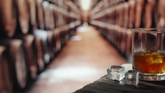 Two glasses of whiskey with ice are on a wooden table. In the background are barrels of whiskey.