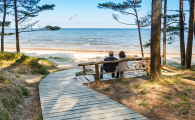 Happy couple of seniors are resting on wooden bench and  looking at the distance on sandy beach of...