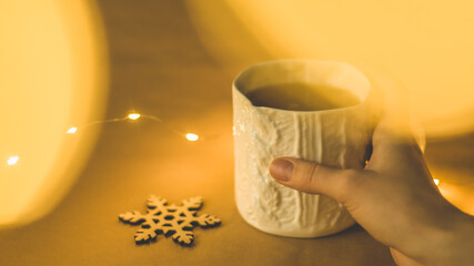 Hand grabbing a beautiful ceramic mug with hot spicy tea in a warm Christmas winter setting with golden lights and a wooden snowflake 