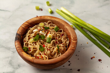 close up image of a wooden bowl full of cooked ramen noodles seasoned with herbs and spices as well as pieces of chopped spring onion. Leftover spring onion, chopped pieces and spices are on marble