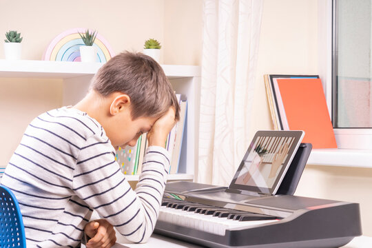 Sad frustrated boy has difficulty learning to play piano at home. Kid using digital tablet to watch video lesson. Online learning difficulties