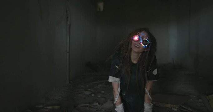 Young woman wearing trendy goggles with a shining light and illuminated frames standing grinning seductively at the camera in the darkness of an abandoned building