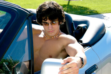 A young hot shirtless and muscular man looks at himself in his rearview side mirror of his sports...