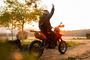 Enduro racer sitting on his motorcycle watching the sunset doing victory sign with hand
travel...