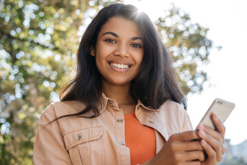 Beautiful smiling African American woman holding mobile phone, shopping online, looking at camera. Closeup portrait of young happy student using smartphone, studying online. Online education concept 