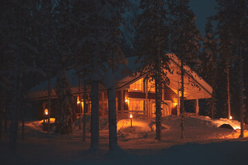 A night view of cozy wooden scandinavian cabin cottage chalet house covered in snow near ski resort in winter with the lights turn on, evening picture