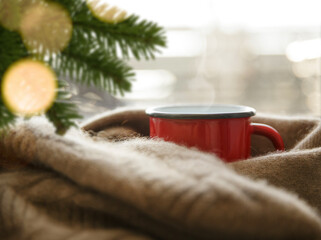 hot drink in a red metal mug wrapped in a cashmere scarf on a cold day under spruce branches