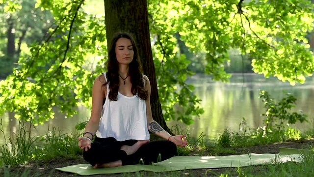 Attractive young, long-haired brunette woman with tattooed arm meditating outdoors in beautiful lush green park. She sits on yoga mat, cross-legged in Comfortable Pose, with tree and lake behind her