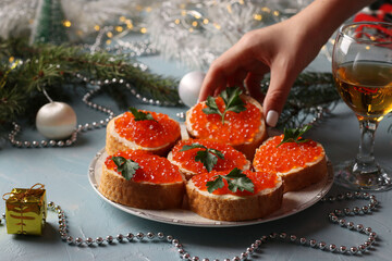 Sandwiches with red caviar on a plate against a light blue background. Festive snack. The woman reaches out to take the sandwich off the plate. - obrazy, fototapety, plakaty